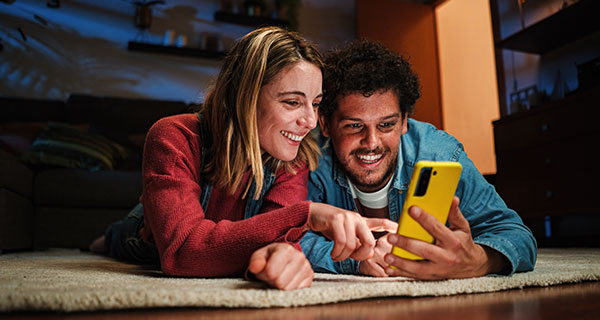 A couple lays on the floor and looks at their phone while smiling. The woman is pointing to something on the phone.  A couple lays on the floor and looks at their phone while smiling. The woman is pointing to something on the phone.